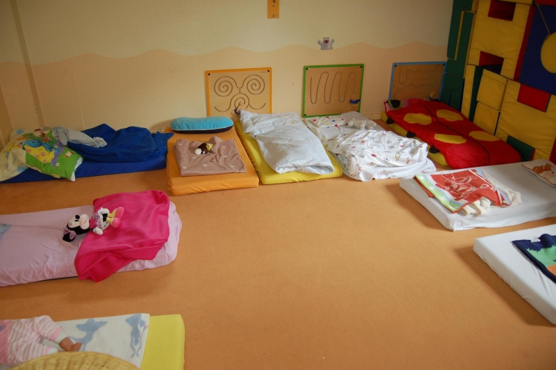 Es sind neun Kindermatratzen zu sehen auf sandfarbenem Teppichboden, U-förmig angeordnet. Die hintere Wand zeigt eine wellenförmige Linie in orange, es sind drei Motoriktafeln darauf angebrac