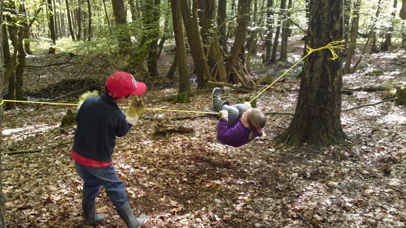  Waldboden mit Laub bedeckt. Ein zwischen zwei Bäumen gespanntes gelbes Seil, an dem ein Kind mit angewinkelten Knien hängt, das von einem weiteren Kind Anschwung bekommt.