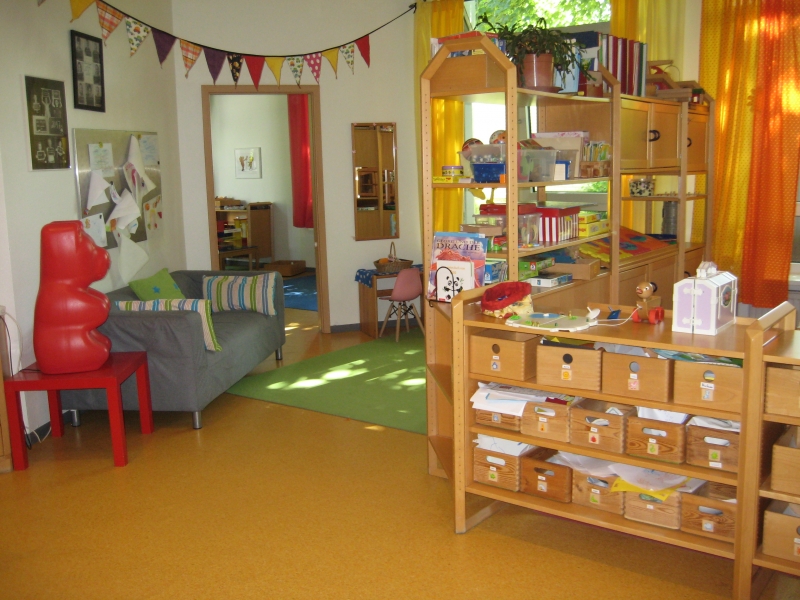 Das Bild zeigt einen Teil eines Gruppenraumes mit Eigentumsfächern der Kinder, Frisierecke, Spielteppich, Sofa und Einblick in die Bauecke.