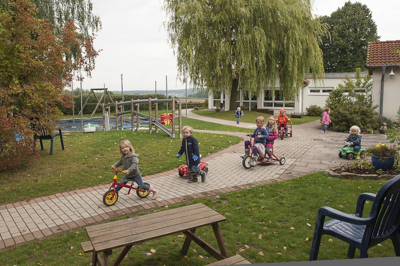 Unser Außengelände bietet vielseitige Möglichkeiten. Hier fahren Kinder mit verschiedenen Fahrzeugen auf befestigten Wegen. Man sieht den großen, runden Sandkasten, den Rasen und eine Schauke