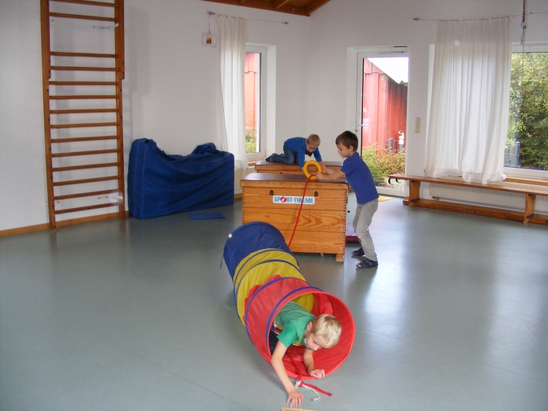 Dies ist unsere große, helle Turnhalle, links an der Wand eine Sprossenwand, im Hintergrund Bänke. Kinder bringen einen Ring über einen Kasten, durch einen Kriechtunnel quer durch die Turnahl