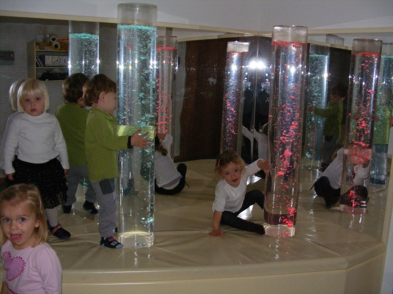 Im Bild ist unser Snoezelen Raum, ein Entspannungsraum. Zu sehen sind Kinder vor 2 großen Farbwechsel-Blasensäulen, dahinter ist eine Spielgelwand. Nicht im Bild ist das Klangwasserbett.