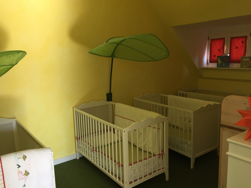 Im Schlafzimmer stehen mehrere Gitterbetten nebeneinander, die mit einem Babynest, Bettwäsche und zum Teil mit einem kleinen Himmel ausgestattet sind.